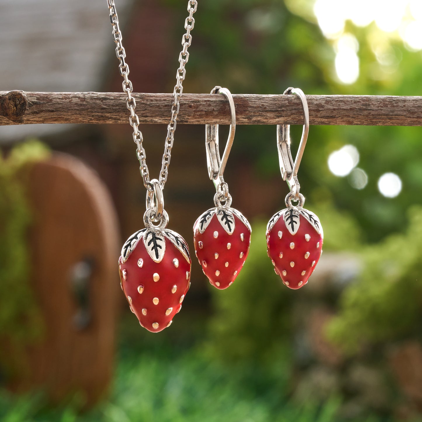Hand-painted red enamel strawberry leverback earrings with matching necklace hanging in garden