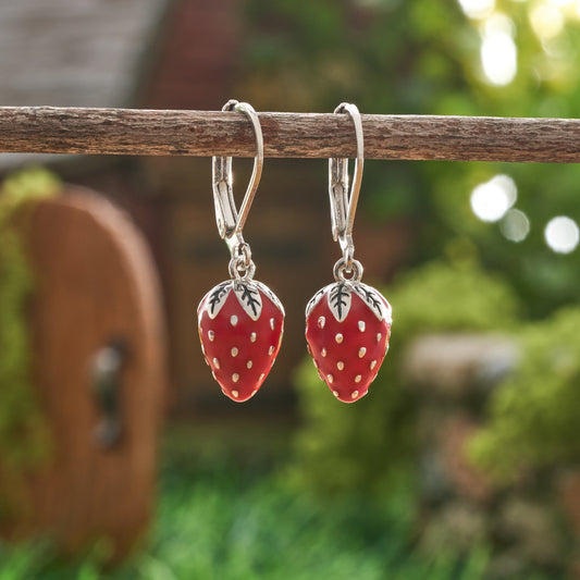 Hand-painted red enamel strawberry leverback earrings hanging from branch in garden setting

