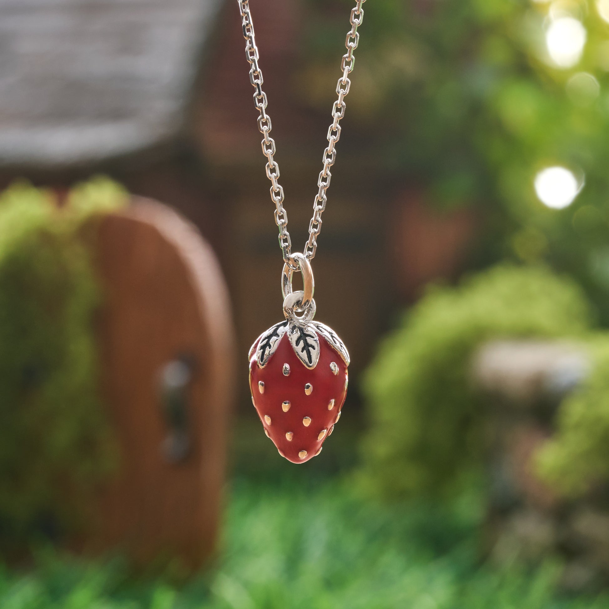 Hand-painted red enamel strawberry necklace front view with silver leaf cap in garden setting