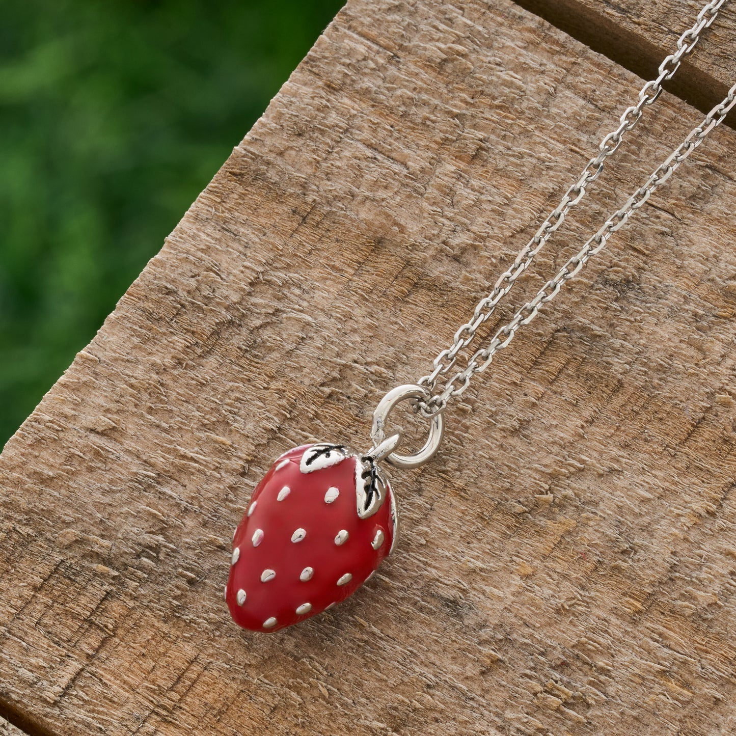 Hand-painted red enamel strawberry necklace side view with silver chain on rustic wood background