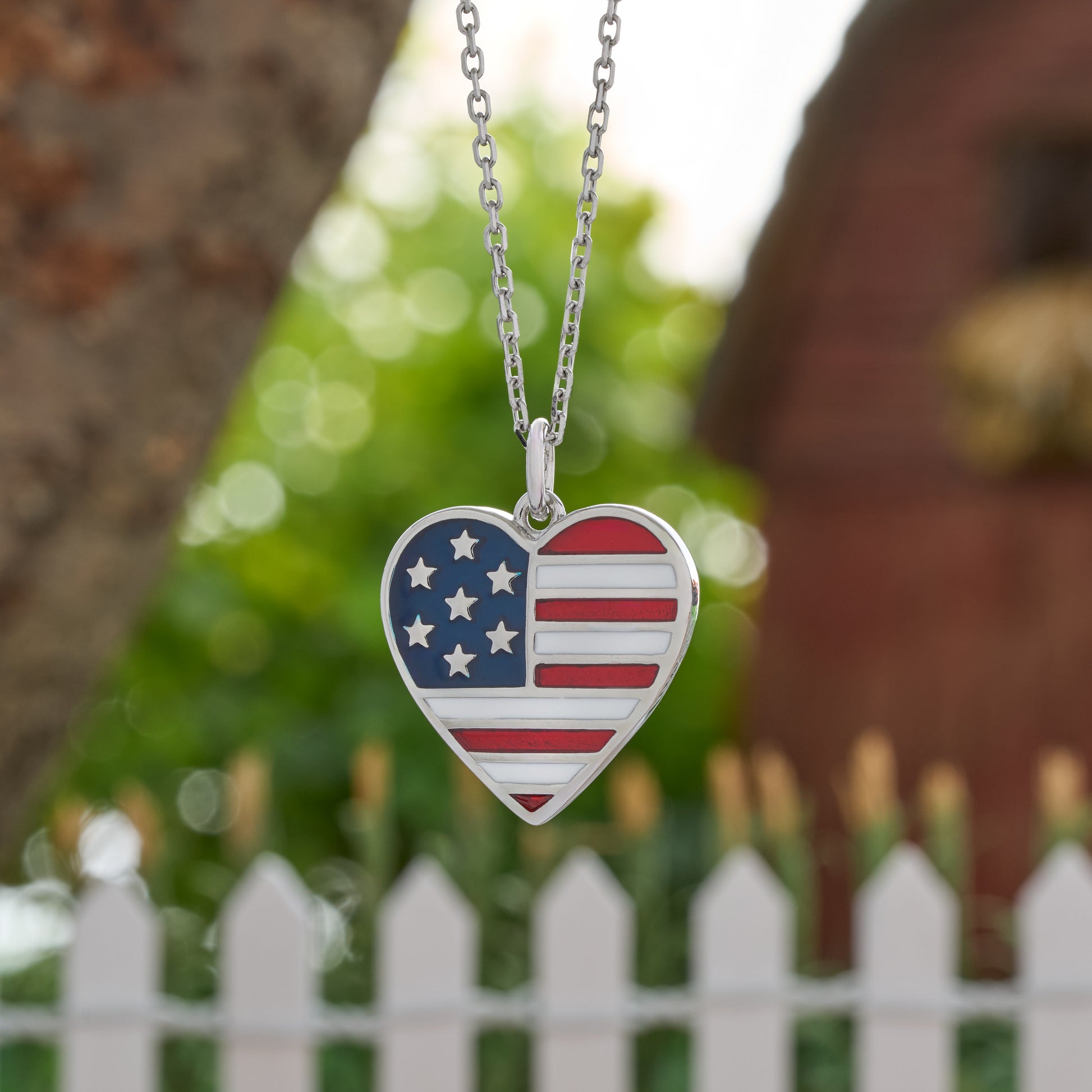 American flag heart pendant necklace with red white and blue enamel and silver chain by white picket fence

